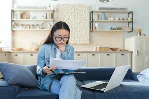 frustrato e triste attività commerciale donna a casa fare carta opera, asiatico donna deluso con opera risultato, utilizzando il computer portatile per calcolare fatture e domestico bilancio foto