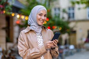 adulto arabo donna indossare pastello hijab e Tenere moderno smartphone mentre avendo camminare su centrale strada. affabile di moda femmina utilizzando mobile applicazione per fabbricazione revisione di visitato ristorante. foto