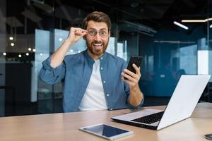 ritratto di un' uomo d'affari soddisfatto con il risultati dentro un' moderno ufficio, un' uomo celebra un' vittoria, sembra a il telecamera detiene un' Telefono nel il suo mani, un' programmatore usi un' il computer portatile a opera. foto