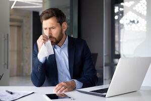 un' malato giovane uomo uomo d'affari si siede nel il ufficio a un' scrivania con un' il computer portatile e documenti, salviette il suo naso con un' tovagliolo, soffre a partire dal un' freddo e un' virus. foto