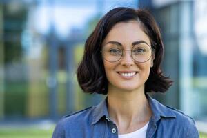 avvicinamento ritratto di un' giovane bellissimo donna nel bicchieri e un' camicia, un' alunno sorridente e guardare a il telecamera al di fuori un' Università città universitaria su un' soleggiato giorno. foto