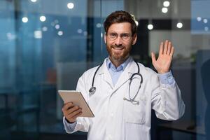 anziano maturo medico con tavoletta guardare a telecamera e Tenere mano su saluto gesto, uomo nel bianca medico cappotto Lavorando dentro moderno clinica, utilizzando tavoletta computer a opera. foto