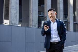 professionale asiatico uomo nel un' elegante completo da uomo sorrisi come lui testi su il suo Telefono, in piedi su un' soleggiato urbano marciapiede. foto