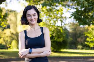 ritratto di un' sorridente e ribelle giovane donna in piedi nel un' parco al di fuori nel nero Abiti, attraversamento sua braccia su sua il petto, sorridente a il telecamera. foto