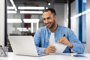 focalizzata indiano uomo Lavorando su un' il computer portatile nel un' contemporaneo ufficio ambiente, assunzione Appunti nel un' occupato giornata di lavoro atmosfera. foto