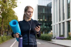 un' focalizzata femmina atleta trasporto un' yoga stuoia e idratazione, ascoltando per musica o un' podcast, e controllo sua smartphone prima un all'aperto fitness sessione nel un' città ambiente. foto