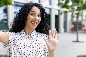 sorridente giovane latino americano donna d'affari nel un' polka punto camicetta agitando ciao, in piedi all'aperto vicino un ufficio. foto