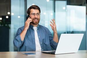 riuscito libero professionista Lavorando dentro moderno ufficio costruzione, uomo parlando su Telefono, uomo d'affari utilizzando il computer portatile a opera foto