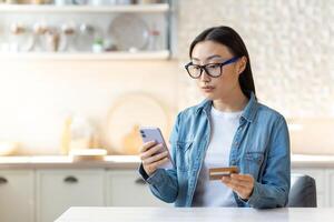 irritato asiatico donna provando per rendere un' Acquista nel un in linea in linea negozio, donna a casa seduta su un' divano nel il cucina utilizzando un applicazione su un' smartphone e Tenere un' banca credito carta foto