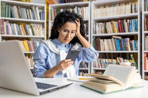 ansioso ispanico alunno guardare a Telefono con il computer portatile e libri a biblioteca scrivania. concetto di fatica nel formazione scolastica e apprendimento le difficoltà. foto