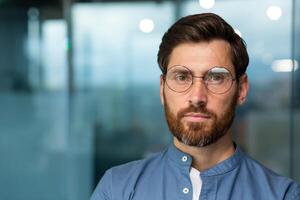 avvicinamento ritratto di grave e riflessivo maturo uomo d'affari con barba, uomo nel camicia e bicchieri guardare a telecamera, startupper Lavorando dentro moderno ufficio costruzione. foto