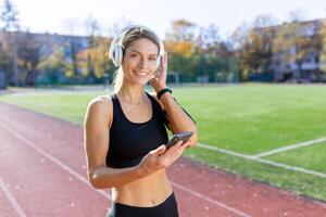 sorridente atletico donna con cuffie la scelta musica su sua smartphone a un' soleggiato traccia campo. concetto di fitness e tecnologia. foto