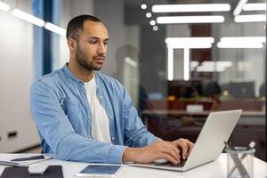 concentrato e grave giovane ispanico uomo Lavorando nel un' moderno ufficio, seduta a un' scrivania e digitando su un' il computer portatile. foto