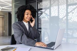 riuscito donna d'affari parlando su il Telefono dentro il ufficio a posto di lavoro, africano americano donna sorridente contento Lavorando utilizzando il computer portatile a opera. foto