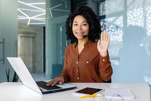 ritratto di in linea cliente servizio chiamata centro lavoratore, donna sorridente e guardare a telecamera, agitando mano gesto di saluto, africano americano donna con cuffia Telefono e il computer portatile per chiamata. foto