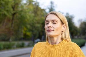 un' di mezza età donna con bionda capelli gode il caldo luce del sole nel un' sereno parco ambientazione, indossare un' accogliente giallo maglione, occhi Chiuso, Abbracciare una persona la tranquillità. foto