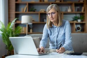 elegante maturo donna seduta su divano e utilizzando portatile computer mentre scrittura nel taccuino a casa. attento femmina unendo a distanza classi per Avanzate formazione e fabbricazione osservazioni a partire dal docente. foto