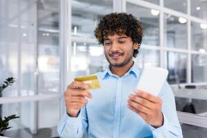 giovane riuscito indiano uomo vicino su nel moderno leggero ufficio interno Tenere banca credito carta e Telefono, uomo d'affari sorridente contento e fabbricazione Acquista nel Internet memorizzare in linea. foto