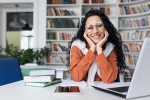 ritratto di contento sorridente indiano femmina insegnante seduta a scrivania nel aula nel davanti di taccuino e guardare a telecamera, conduzione in linea classi, giovane riuscito tutore, in linea alunno. foto