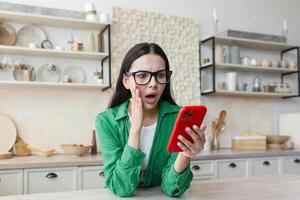 un' preoccupato e scioccato giovane donna è in piedi a casa nel il cucina, Tenere il Telefono nel sua mani foto