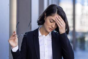 giovane donna stanco con male alla testa, donna d'affari al di fuori ufficio edificio strofina occhi, dare le vertigini, oberati di lavoro lavoratore all'aperto nel attività commerciale completo da uomo vicino su. foto