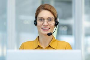 ritratto di maturo femmina cliente servizio lavoratore, maturo attività commerciale donna con chiamata cuffia sorridente e guardare a telecamera, chiamata centro in linea cliente servizio, dentro ufficio a posto di lavoro. foto