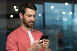 sorridente e contento uomo d'affari dentro il ufficio di il finestra usi il Telefono un' maturo uomo con un' barba sembra a il smartphone schermo, sfoglia Internet pagine, tipi messaggi nel un in linea applicazione foto