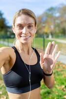 femmina atleta dopo jogging nel stadio parlando con amici e registrazione in linea blog, maturo bionda con cuffie guardare a smartphone telecamera sorridente, chiamata con cuffia. foto