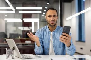 grave e preoccupato giovane uomo seduta nel il ufficio a il scrivania, Tenere un' mobile Telefono nel il suo mano e guardare deluso a il telecamera mentre diffusione il suo mani. foto