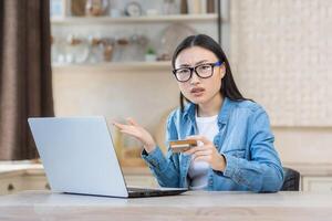 ritratto di insoddisfatto e arrabbiato imbrogliato donna, asiatico donna a casa nel cucina provando per rendere banca trasferimento e Acquista nel in linea Internet negozio, giovane casalinga utilizzando il computer portatile guardare a telecamera foto