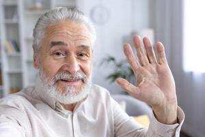 ritratto di un' allegro anziano uomo con un' barba, agitando per il telecamera a partire dal un' accogliente, pieno di luce vivente spazio. foto