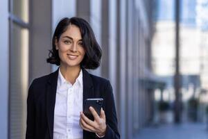 avvicinamento foto di un' giovane femmina avvocato nel un' attività commerciale completo da uomo in piedi vicino un' tribunale, Tenere un' Telefono e sorridente a il telecamera.