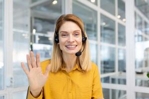 ritratto di un' sorridente giovane donna d'affari seduta nel il ufficio nel un' cuffia e parlando per il telecamera. conduce un' attività commerciale incontro, saluta e onde a il telecamera. foto