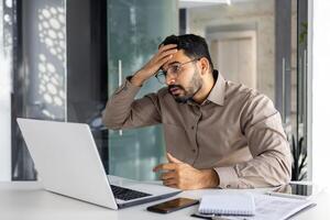 irritato e deluso uomo d'affari a posto di lavoro avuto negativo finanziario risultati, insoddisfatto uomo Lavorando dentro ufficio a posto di lavoro con il computer portatile. foto