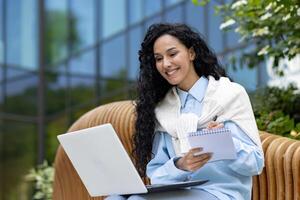 giovane femmina alunno studiando al di fuori Università città universitaria, latino americano donna con il computer portatile seduta su panchina e registrazione classe conferenza, contento sorridente. foto