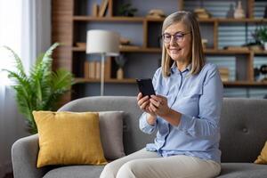 anziano dai capelli grigi donna seduta con gioia su divano a casa, pensionato Tenere Telefono nel mani, utilizzando App su smartphone, digitando testo Messaggio, chiamata, e navigazione sociale reti con un' Sorridi. foto