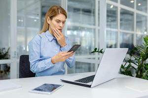 ha riguardato professionale femmina nel un' blu camicia a sua ufficio scrivania guardare perplesso a sua smartphone con un' il computer portatile e tavoletta qui vicino. foto