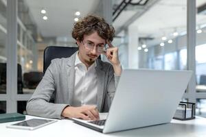 grave giovane maschio professionale con Riccio capelli si concentra su il suo opera a un' il computer portatile nel un' ben illuminato ufficio ambientazione. foto