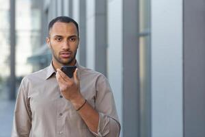 arrabbiato uomo d'affari nel camicia registrazione Audio Messaggio a partire dal al di fuori ufficio costruzione, africano americano uomo utilizzando assistente dando comandi per artificiale intelligenza utilizzando smartphone. foto