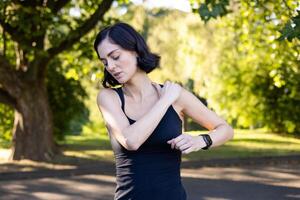 un' avvicinamento foto di un' giovane donna fare gli sport nel il parco, in piedi e Tenere sua mano di sua spalla, tirato un' muscolo, si sente acuto dolore, è fare un' massaggio.