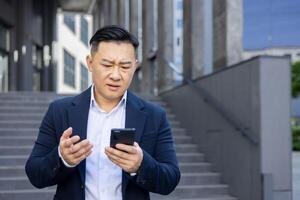 avvicinamento foto di un' preoccupato giovane asiatico uomo d'affari uomo nel un' completo da uomo in piedi al di fuori e guardare irritato a il Telefono, lui è Tenere nel il suo mani.