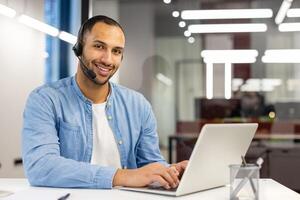 fiducioso maschio cliente supporto agente indossare un' cuffia mentre Lavorando su il computer portatile nel un' luminosa moderno ufficio ambientazione. foto