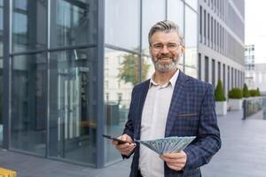 contento capo in piedi su il strada con un' pila di dollari nel il suo mani e un' Telefono, vincente il lotteria, successo a opera, sorridente e guardare a il telecamera. foto