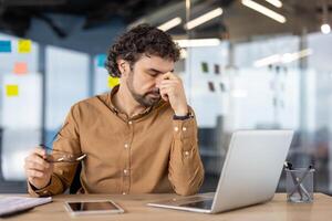 stanco e sopraffatto uomo d'affari ha vertigini e occhio dolore, maturo esperto maschio lavoratore a posto di lavoro dentro ufficio, Lavorando con il computer portatile. foto