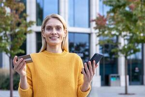 fiducioso biondo donna nel giallo maglione con gioia Tenere un' credito carta e smartphone, in piedi all'aperto con un' moderno edificio sfondo. foto