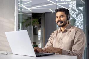 un' professionale uomo con un' cuffia utilizzando un' il computer portatile nel un' moderno ufficio ambientazione, veicolare cliente servizio e tecnologia. foto