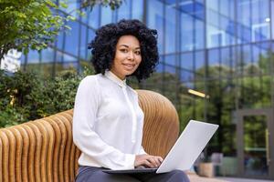 ritratto di un' giovane africano americano donna d'affari seduta al di fuori un ufficio centro su un' panchina e utilizzando un' il computer portatile. lui sorrisi e sembra compiaciuto in il telecamera. foto