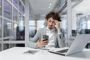 giovane adulto maschio ufficio lavoratore sensazione stressato e oberati di lavoro come lui offerte con multitasking e scadenze, guardare a il suo Telefono con un' preoccupato espressione. foto