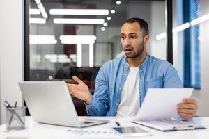 irritato giovane musulmano uomo seduta nel moderno ufficio a scrivania, Tenere documenti nel mani e guardare a il computer portatile schermo nel shock, diffusione mani. foto