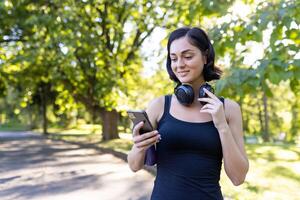 avvicinamento foto di un' giovane ragazza jogging e fare gli sport nel il parco, in piedi nel cuffie e utilizzando un' mobile Telefono.